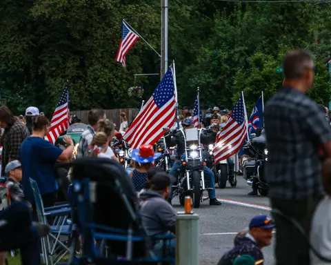 Motorcyclists carrying American flags ride down a street past a crowd of spectators.