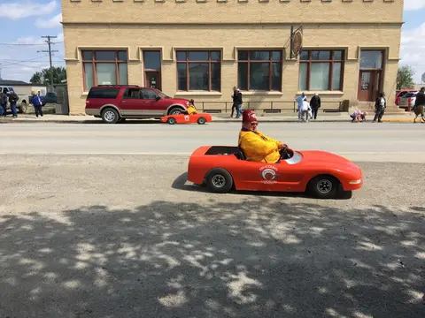A person drives a small red car on a street in front of a brick building.