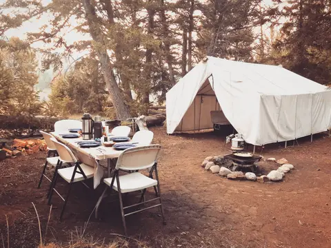 A campsite with a large white tent, a set-up dining table, and a fire pit in a wooded area.