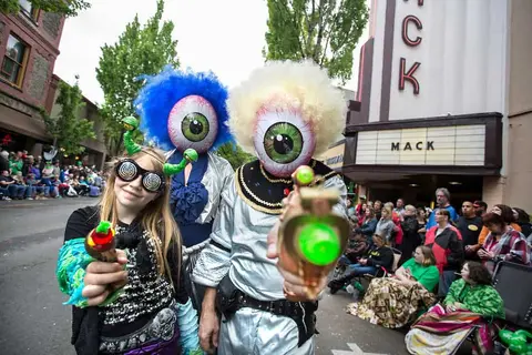 People in costumes with giant eyeball heads and toy ray guns pose outside near a crowd.