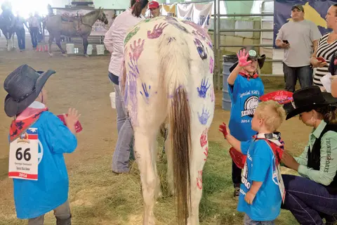 Children put painted handprints on a horse at an indoor arena event.