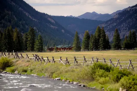 A scenic mountain valley with a river, a wooden fence, and cabins.
