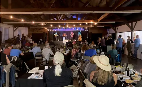People seated at tables inside a rustic hall watching musicians perform on a stage.