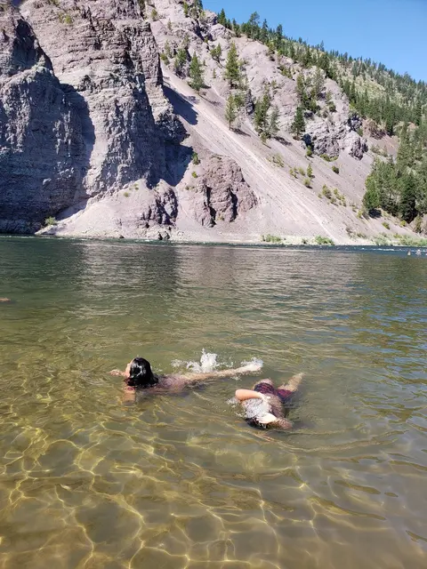 Two people swimming in a river with rocky cliffs in the background.