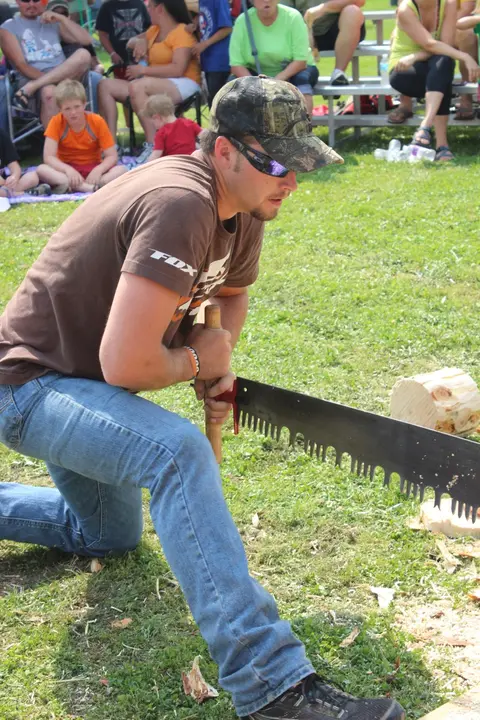 A man kneels on the grass and uses a large saw to cut a log.