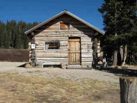 A rustic wooden cabin stands in a clearing near a forest under a clear blue sky.