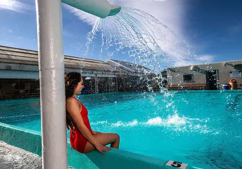 A woman sits at the edge of a swimming pool beneath a water feature.