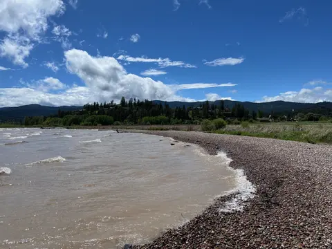 A pebbled shoreline along a body of water with rolling hills under a blue sky.