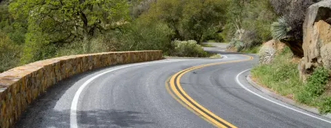A winding asphalt road curves through a landscape with trees and a stone wall.