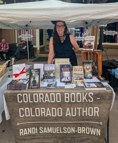 An author stands behind her table displaying Colorado-themed books at an outdoor market.