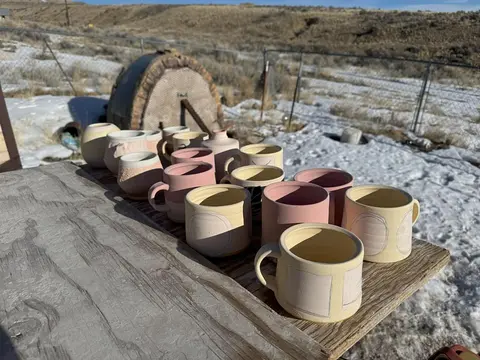A collection of ceramic mugs and pots sits on a wooden table outdoors in a snowy landscape.