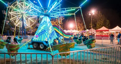 A carnival ride operates at night with lights and a ferris wheel in the background.