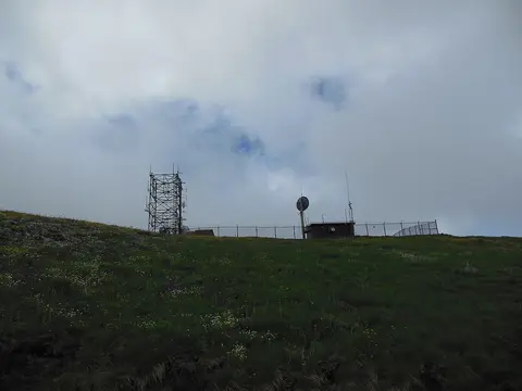 A communication tower and building stand on a grassy hilltop under a cloudy sky.