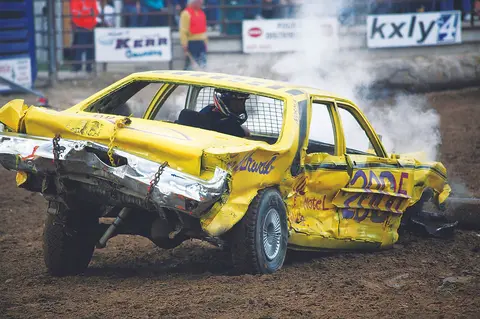 A heavily damaged yellow car smokes in a dirt arena during a demolition derby.