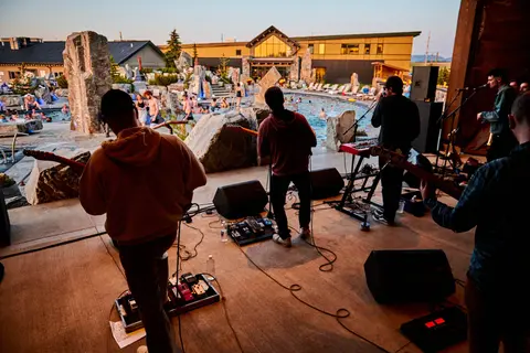 A band performs on a stage outdoors with a pool and resort buildings in the background.