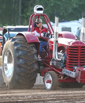 A person sitting on a red tractor with a helmet on the roll bar.