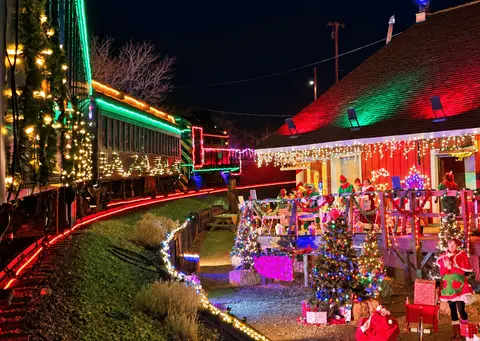 A decorated holiday train passes by a lit-up building at night.