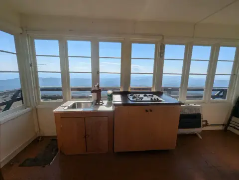 An interior view of a room with large windows showing a scenic view and a small kitchen setup.