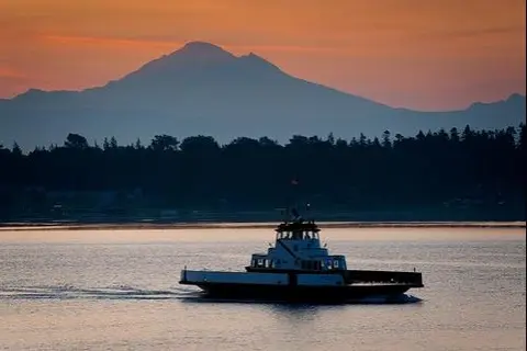 A ferry travels on the water with a mountain and sunset sky in the background.