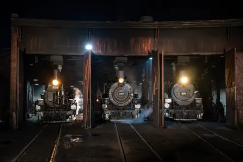 Three steam locomotives sit inside an engine house at night with their headlights on.