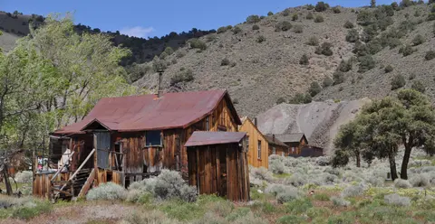 Old wooden buildings with rusted metal roofs stand in a dry mountainous landscape.