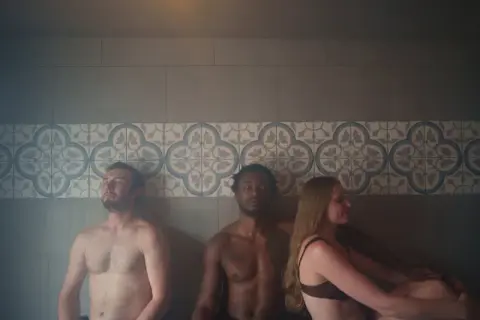 Three people relax inside a tiled steam room.