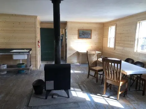 An interior view of a room with wood paneling walls, a wood-burning stove, a table, and chairs.