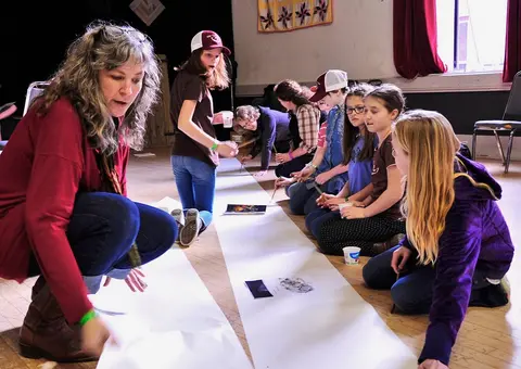 An instructor and children work on a long paper scroll indoors.
