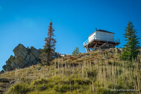 A small cabin sits on a rocky ridge under a clear blue sky.