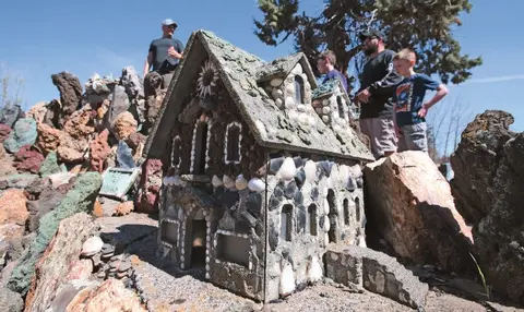 A model house decorated with rocks and shells stands outdoors with people nearby.