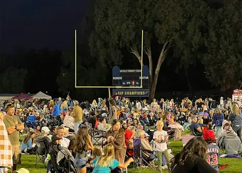A large crowd gathered on a grassy field at night in front of a scoreboard and goalpost.