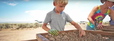 A young boy sifting through gravel in a wooden screen box outdoors.