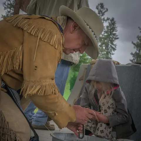 A man in fringe buckskin and a hat interacts with a young child outdoors.