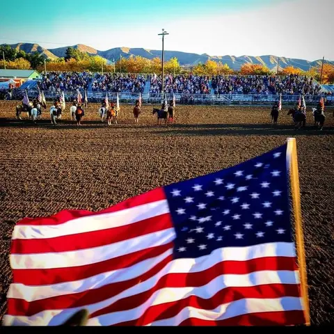 An American flag is held in the foreground at a rodeo arena with riders and a crowd.