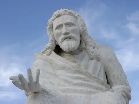 A large white stone statue of a man with a beard and outstretched hand against a blue sky.
