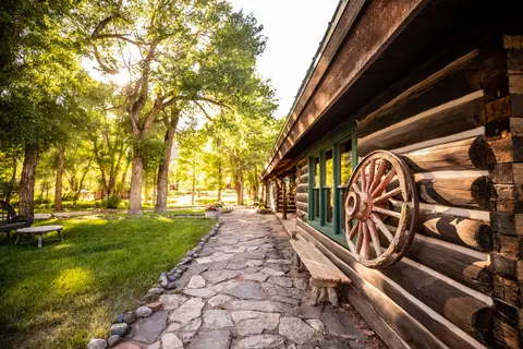 A stone pathway runs alongside a rustic log building with a wagon wheel mounted on the wall.