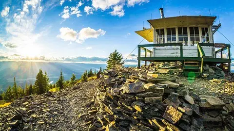 A fire lookout building sits atop a rocky mountain summit during sunset.