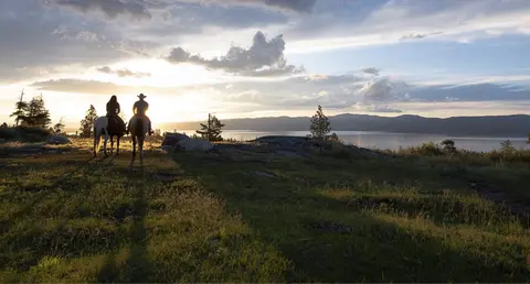 Two people on horseback look out over a large lake at sunset.