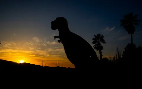 A dinosaur statue is silhouetted against a sunset sky.