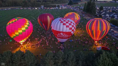 Aerial view of several hot air balloons glowing on a grassy field at dusk with a crowd of people gathered around.