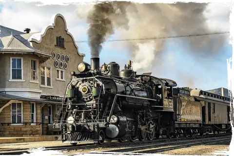A steam locomotive emits smoke while stopped next to a historic station building.