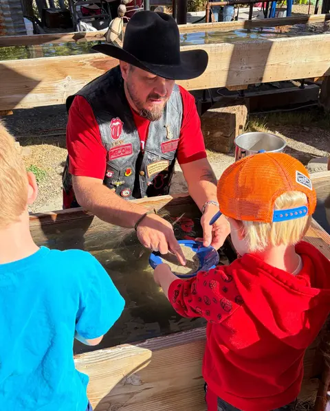 A man in a cowboy hat helps children pan for gold at a water trough.