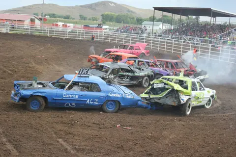 Several damaged cars are involved in a demolition derby in a dirt arena with spectators in the stands.