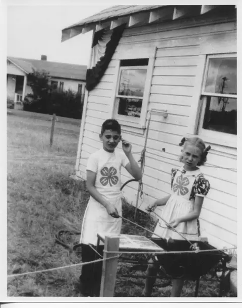 Black and white historic photo of two children standing outside a building by a grill.