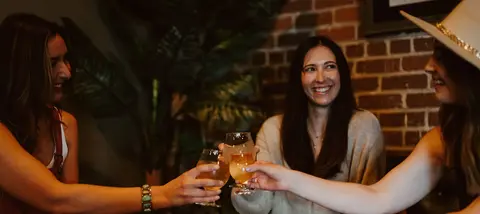 Three women toast glasses of drinks together indoors.