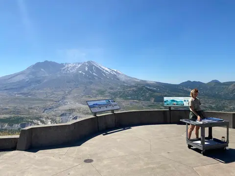A park ranger stands by a cart on an observation deck overlooking a large mountain.