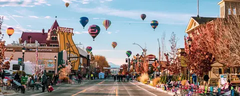 Hot air balloons fly over a crowded city street with buildings and autumn trees.