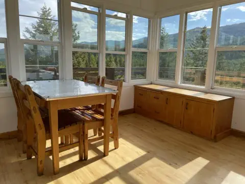 A brightly lit sunroom features a wooden dining table and chairs with scenic views through large windows.