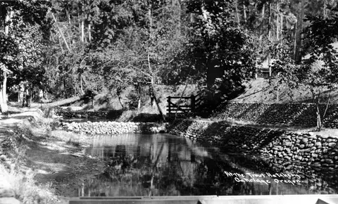 A historic black and white photograph of a stream with stone walls and surrounding trees.