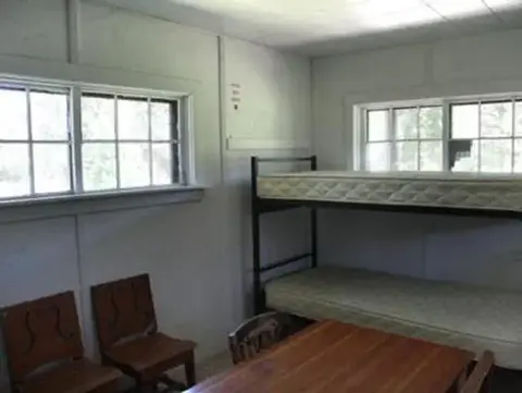Interior view of a simple room with a bunk bed, table, and chairs near windows.